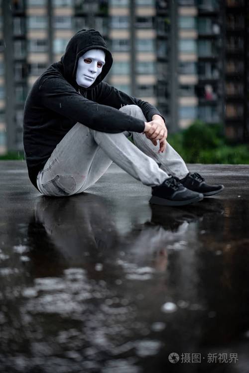 男人在白色面具下拥抱着他的膝盖坐在废弃建筑的屋顶上的雨中双相情感